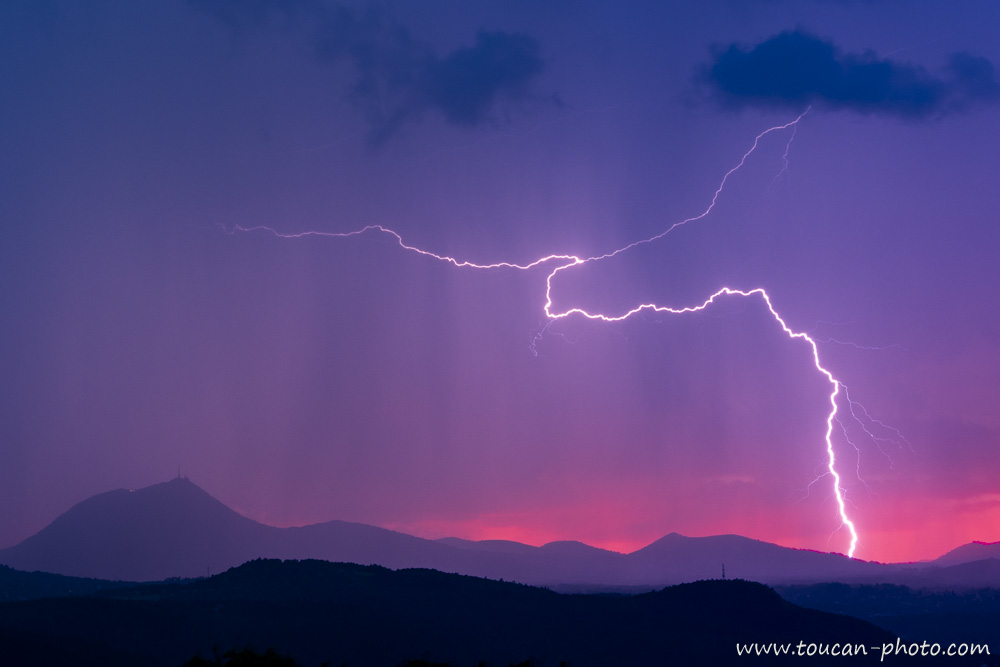 Foudre frappant près du puy de Dôme au coucher du soleil, avec un ciel dramatique violet et rouge au-dessus d’un paysage montagneux en France
